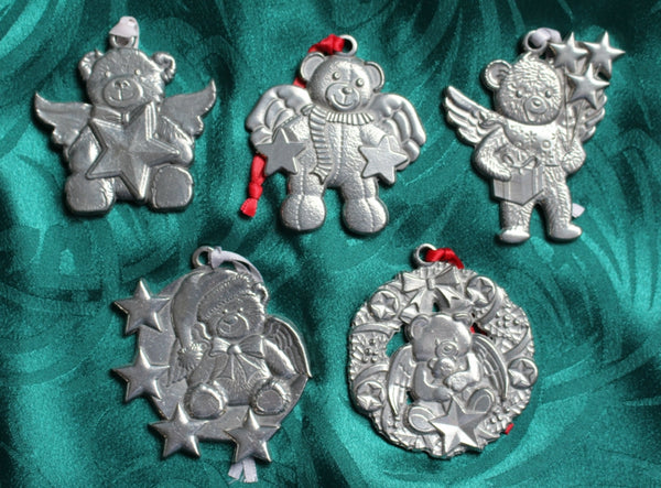 Five pewter ornaments, each featuring a bear, angel wings, and stars, laid on green silk.