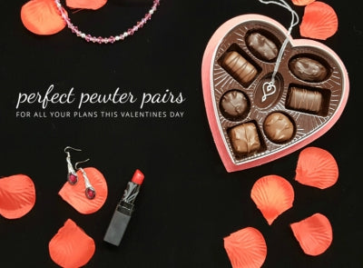 a black background is covered with red silk flower petals. a pewter heart pendant is inside a box of chocolates and a pair of pewter earrings is beside a red lipstick. text reads "perfect pewter paitings for all your plans this valentines day"